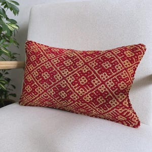 Op de afbeelding: Een rechthoekig sierkussen van geweven stof met een herhalend geometrisch ruitpatroon in donkerrood en beige. Het textiel heeft een getextureerd uiterlijk met zichtbare verweven draden die complexe vormen over het oppervlak vormen.