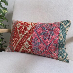 Op de afbeelding: Een rechthoekig sierkussen met een geweven textielhoes met een veelkleurig geometrisch patroon. Het ontwerp bevat secties in donkerrood, felroze en gedempt groenblauw, geaccentueerd met crèmekleurige ruit- en bloemmotieven.