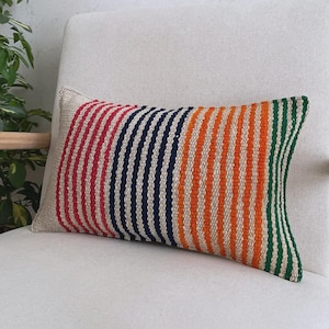 Op de afbeelding: Een rechthoekig sierkussen met een geweven textuur en verticale strepen in rood, marineblauw, oranje en groen op een natuurlijke beige achtergrond. De stof heeft een zichtbaar, grof weefpatroon over het gehele oppervlak van het kussen.