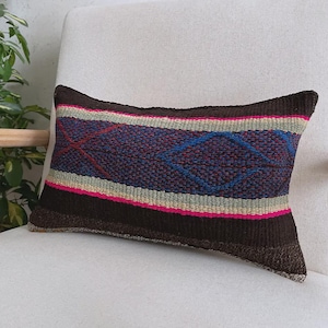 Op de afbeelding: Een rechthoekig sierkussen met een geweven textuur en horizontale strepen in donkerbruin, felroze en lichtbeige. Het middenpaneel heeft een donkerblauwe achtergrond met een geometrisch ruitpatroon omlijnd met rode en blauwe draad.