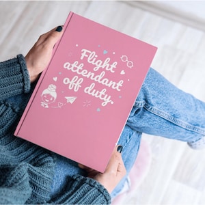 May include: A pink journal with the text "Flight attendant off duty" in white. The cover features a cartoon illustration of a flight attendant, hearts, and a paper airplane. The journal is held by a person wearing a blue sweater and jeans.
