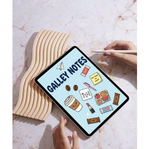 May include: A tablet displaying "GALLEY NOTES" with various illustrated icons, including coffee, lipstick, and a lunchbox. A wooden, wavy decorative piece is to the left. A hand holds a stylus, and another hand is visible at the bottom.