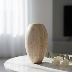 May include: A beige, oval vase with a textured surface. The vase is made of a light-coloured stone and sits on a white marble table. The table has black legs. Decorative elements are on the table.