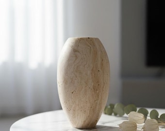 Puede incluir: Un jarrón beige de forma ovalada con una superficie texturizada. El jarrón está hecho de piedra de color claro y se encuentra sobre una mesa de mármol blanco. La mesa tiene patas negras. Hay elementos decorativos sobre la mesa.