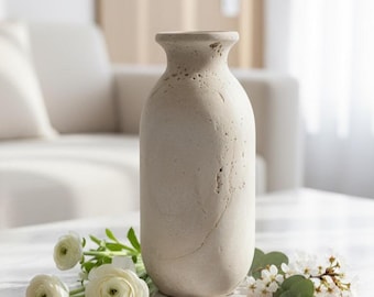 Puede incluir: Un jarrón beige texturizado con cuello estrecho y cuerpo redondeado. Se encuentra sobre una mesa de mármol blanco, acompañado de flores blancas y vegetación. El fondo presenta un sofá y una pared de tonos neutros.