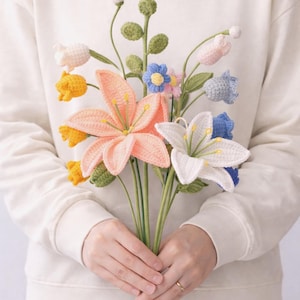 Könnte beinhalten: Ein Strauß aus handgefertigten Häkelblumen mit einer großen pfirsichfarbenen Lilie, einer weißen Lilie, gelben glockenförmigen Blüten, kleinen blauen und rosa Blumen sowie grünen Stielen mit Blättern. Die Blumen zeigen deutlich sichtbare Maschen.