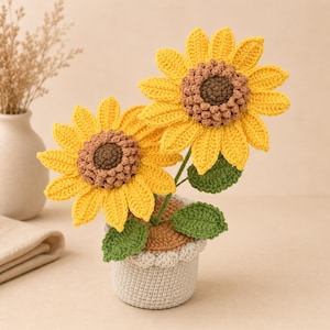 Puede incluir: Dos girasoles de ganchillo con pétalos de color amarillo brillante y centros marrones texturizados, con hojas verdes en los tallos. Las flores están plantadas en una pequeña maceta cilíndrica de ganchillo gris claro con borde superior beige.