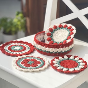 Può includere: Quattro sottobicchieri all'uncinetto in rosso, bianco e verde con un motivo floreale. I sottobicchieri sono su un tavolo bianco con un cestino all'uncinetto sullo sfondo.