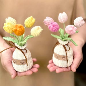 Op de afbeelding: Twee kleine, gehaakte tulpenboeketten in gebroken witte vazen. Eén boeket heeft gele en oranje tulpen, de andere roze. Elke vaas heeft een bruin leren bandje en een label met "Hand Made" en een smiley.