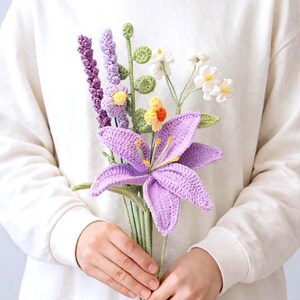 Könnte beinhalten: Ein Strauß handgefertigter Häkelblumen, darunter eine große lila Lilie, Lavendel und weiße Blüten. Die Blumen werden in den Händen einer Person gehalten, mit einem weißen Pullover und blauen Jeans im Hintergrund.