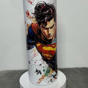May include: White stainless steel tumbler with a colourful abstract design featuring a superhero with a red cape and a yellow symbol on their chest.