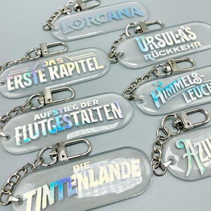 Könnte beinhalten: Ein Set mit acht transparenten Schlüsselanhängern mit schimmerndem Text. Die Anhänger zeigen Titel aus einer Fantasy-Serie, darunter "Lorgana", "Das Erste Kapitel", "Aufstieg Der Flutigestaalten", "Die Tintenlande", "Ursulas Ruckkehr", "Himmels-Leuch" und "Azur".
