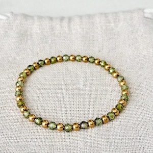 Könnte beinhalten: Ein Armband mit abwechselnd goldenen und grünen Perlen. Das Armband besteht aus kleinen, runden Perlen, die auf einem Gummiband aufgereiht sind. Die grünen Perlen haben ein facettiertes Aussehen, während die goldenen Perlen glatt sind.