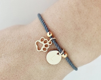 May include: Gray rope bracelet with a gold paw print charm and a gold initial charm. The initial charm is the letter L.