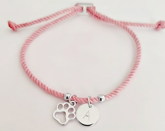 May include: A pink braided cord bracelet with a silver paw print charm and a silver charm with the letter A.