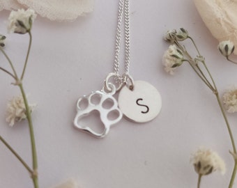 May include: A silver necklace with a paw print charm and a small round charm with the letter 'S' on it. The necklace is on a white background with white flower petals.