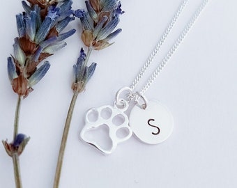May include: Silver necklace with a paw print charm and a small circle charm with the letter 'S' on it. The necklace is displayed on a white background with lavender sprigs.