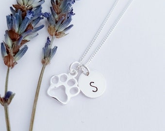 May include: Silver necklace with a paw print charm and a small disc charm with the letter 'S' engraved on it. The necklace is displayed on a white background with lavender sprigs.