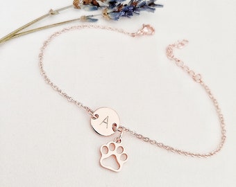 May include: A rose gold chain bracelet with a small round charm engraved with the letter "A" and a paw print charm.