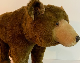 Puede incluir: Un juguete vintage de peluche marrón en forma de oso sobre ruedas. El oso tiene un cuerpo de peluche marrón, una cara de color canela y ojos negros. Las ruedas son rojas y de madera.