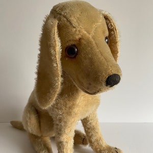 Puede incluir: Un perro de peluche vintage con pelaje marrón, ojos negros y nariz negra. El perro está sentado con las patas metidas debajo.