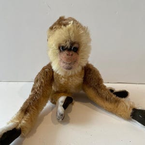 Pode incluir: Um macaco de pelúcia com pelo castanho claro e creme. O macaco tem o rosto e o peito creme, com pelo castanho nos braços e nas costas. As mãos e os pés do macaco são pretos.