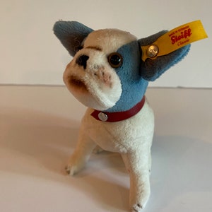 May include: A white and blue plush French Bulldog puppy with brown eyes and a red collar. The puppy is sitting on a white surface and has a Steiff Classic tag attached to its ear. The tag is yellow with black text that reads "Made in Germany Steiff Classic".