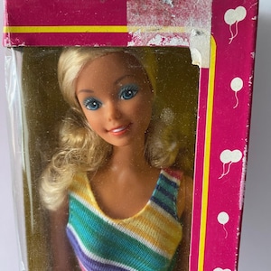 Puede incluir: Una muñeca vintage con cabello rubio y ojos azules, vestida con un top a rayas de colores y una falda blanca. La muñeca está en su embalaje original, que es rosa con gráficos de globos blancos. La caja tiene texto que dice "NO RECOMENDADO PARA NIÑOS MENORES DE 3 AÑOS."