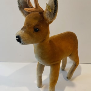 May include: A vintage plush deer toy with brown fur, black eyes, and tan antlers. The deer has a black nose and a small yellow tag on its ear. The toy is standing on four legs.