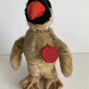 May include: A tan and black stuffed bird with a red beak and a red tag that says "Steiff Tricky Patent ANG".
