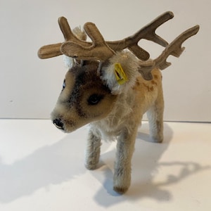 May include: A vintage stuffed reindeer toy with tan antlers and a brown and tan body. The toy has a yellow tag on its ear and is standing on four legs. The reindeer's face is detailed with black eyes and a black nose.