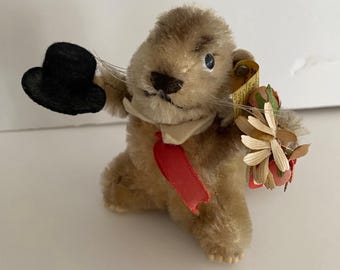 Puede incluir: Una figura de peluche marrón y difusa que sostiene un pequeño sombrero de copa negro en una pata y un ramo de flores artificiales en la otra. La figura lleva un cuello blanco y una corbata roja, con una etiqueta amarilla unida a un lado.