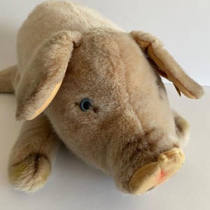 May include: A plush brown pig toy with a blue eye and a pink snout. The pig is lying on its belly with its legs outstretched.