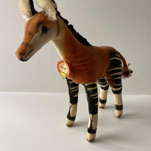 May include: A brown and black stuffed okapi with a white tag that says "Okapi" on it. The okapi is standing on a white surface.