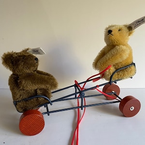 Puede incluir: Dos osos de peluche, uno marrón y otro beige, en un carro de juguete de metal negro con ruedas rojas y una cuerda roja. Los osos llevan ambos etiquetas que dicen "Steiff" y "Hecho en Alemania".