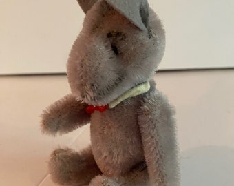 Puede incluir: Un pequeño conejo de peluche gris con una pajarita roja y un collar blanco. El conejo está sentado sobre una superficie blanca.