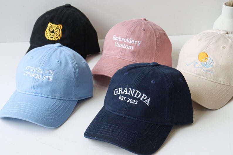 May include: Five different embroidered baseball caps. One is black with a yellow tiger, one is light blue with white coordinates, one is pink with white text that says "Embroidery Custom", one is beige with a yellow sun and a seashell, and one is navy blue with white text that says "GRANDPA EST. 2025".