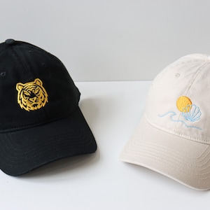 May include: Two baseball caps, one black with a gold embroidered tiger face and one beige with a yellow sun and blue seashell embroidery.