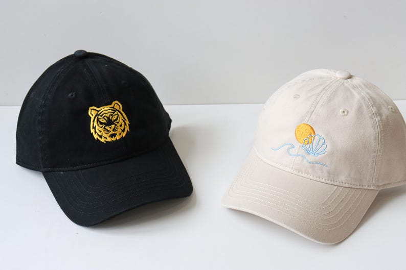 May include: Two baseball caps, one black with a gold embroidered tiger face and one beige with a yellow sun and blue seashell embroidery.