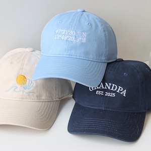 May include: Three baseball caps, one light blue with the coordinates "47&deg;21'30.7"N 12&deg;48'29.3"W" embroidered on the front, one beige with a sun and seashell design, and one dark blue with "GRANDPA EST. 2025" embroidered on the front.