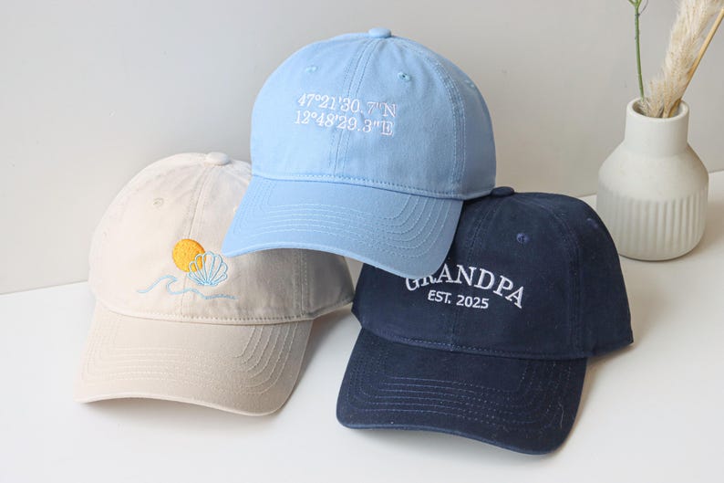 May include: Three baseball caps, one light blue with the coordinates "47&deg;21'30.7"N 12&deg;48'29.3"W" embroidered on the front, one beige with a sun and seashell design, and one dark blue with "GRANDPA EST. 2025" embroidered on the front.
