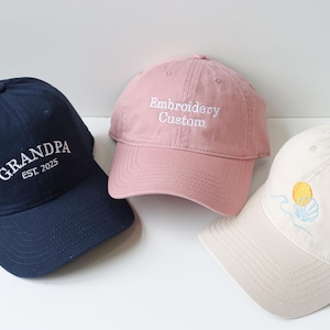 May include: Three embroidered baseball caps. One is navy blue with white embroidery that says "GRANDPA EST. 2025". One is pink with white embroidery that says "Embroidery Custom". One is cream with a yellow and blue embroidered sun and shell design.