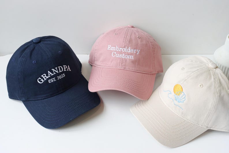 May include: Three embroidered baseball caps. One is navy blue with white embroidery that says "GRANDPA EST. 2025". One is pink with white embroidery that says "Embroidery Custom". One is cream with a yellow and blue embroidered sun and shell design.