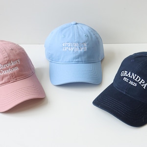 May include: Three baseball caps with embroidered text. The first cap is pink with "Embroidery Custom" in white text. The second cap is light blue with coordinates in white text. The third cap is navy blue with "GRANDPA EST. 2025" in white text.