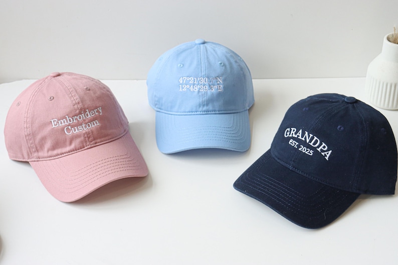 May include: Three baseball caps with embroidered text. The first cap is pink with "Embroidery Custom" in white text. The second cap is light blue with coordinates in white text. The third cap is navy blue with "GRANDPA EST. 2025" in white text.