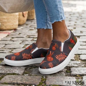 May include: A pair of slip-on sneakers featuring a dark charcoal background with a repeating pattern of orange-red mushrooms and fern-like leaves. The shoes have white rubber soles and white elastic side panels for a secure fit on the feet.