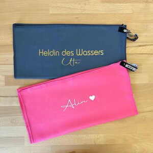 Pu&ograve; includere: Due asciugamani in microfibra, uno grigio con testo dorato che dice "Heldin des Wassers Ute" e uno rosa con testo bianco che dice "Alin" con un simbolo di cuore.