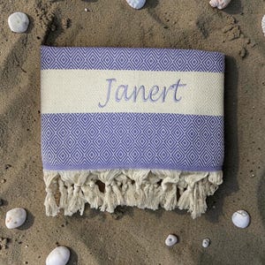 May include: A folded beach towel with a lavender diamond pattern and a cream-colored stripe embroidered with the name "Janert". The towel has cream-colored fringe and is surrounded by seashells and pebbles on sand.