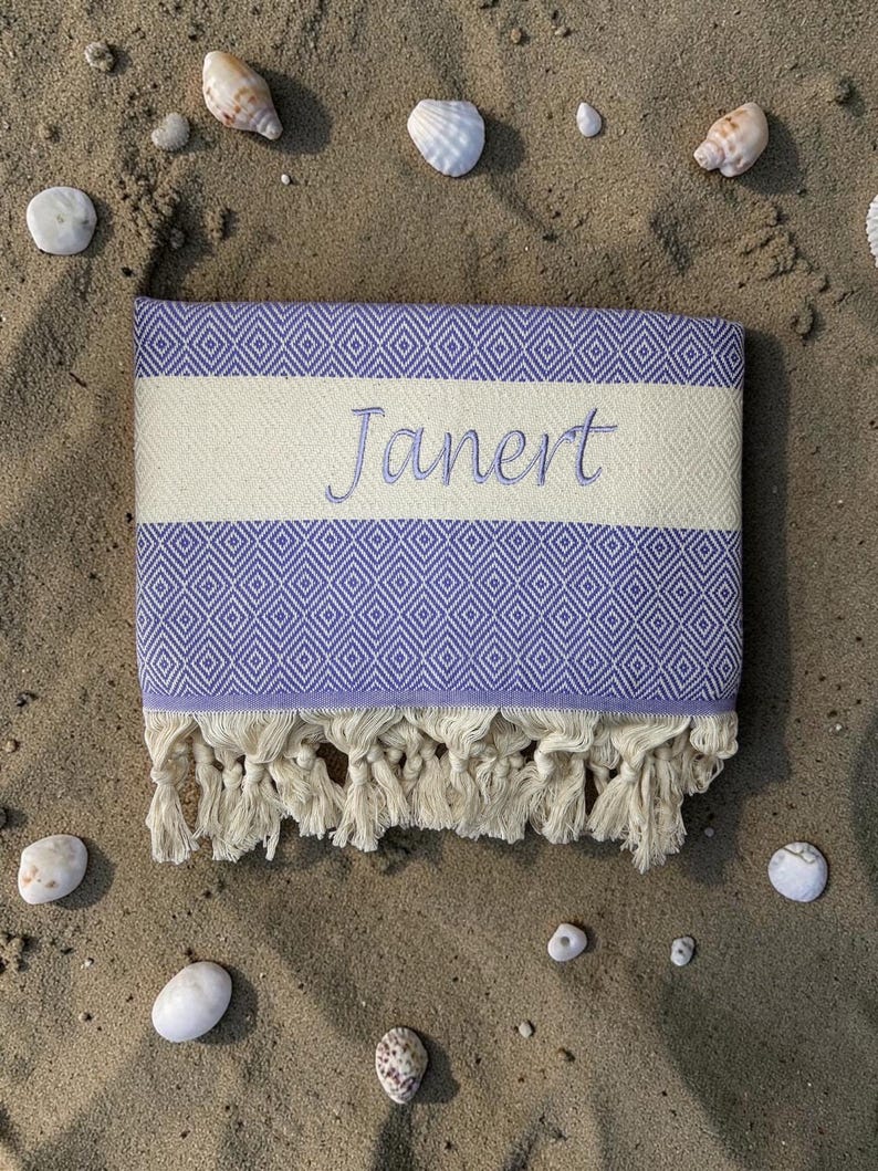 May include: A folded beach towel with a lavender diamond pattern and a cream-colored stripe embroidered with the name "Janert". The towel has cream-colored fringe and is surrounded by seashells and pebbles on sand.