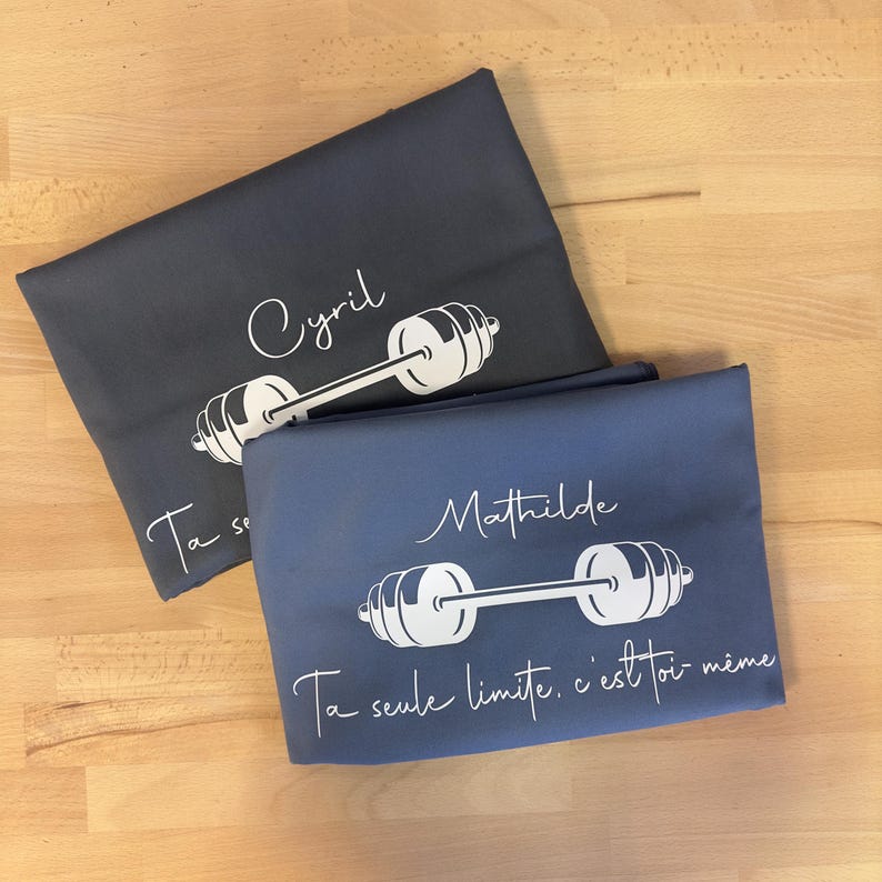 May include: Two folded t-shirts in dark gray and slate blue. Each shirt features a white graphic of a barbell with a name above it, Cyril on the gray shirt and Mathilde on the blue shirt, and the French text Ta seule limite, c'est toi-m&ecirc;me.